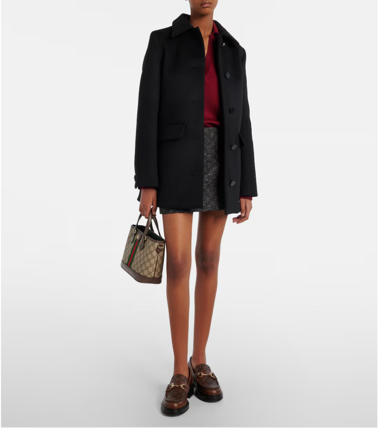 Person wearing a black coat, red shirt, and patterned skirt holding a Gucci handbag on a white background