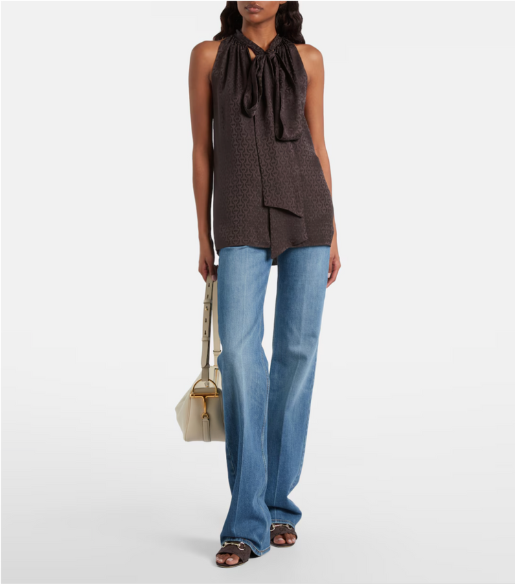 Woman wearing a brown sleeveless top with a tie detail and blue jeans, holding a beige handbag on a white background.