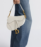 White handbag with gold accents held by a person wearing blue jeans on a light gray background