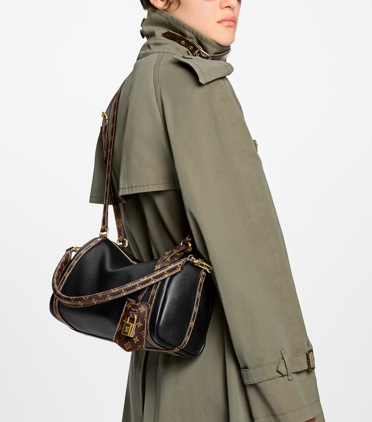 Person wearing a green trench coat holding a black handbag with gold accents on a white background