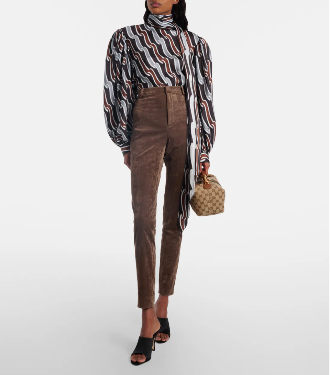 Person wearing a patterned blouse and brown pants holding a Gucci handbag and wearing black gucci mules on a white background