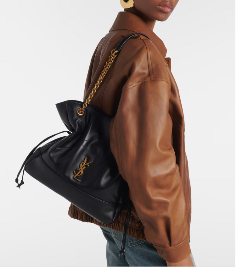 Person wearing a brown leather jacket with a black drawstring bag featuring a gold logo.