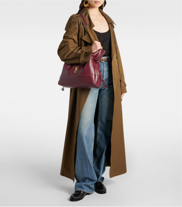 Person wearing a long brown coat, blue jeans, and holding a maroon handbag on a white background