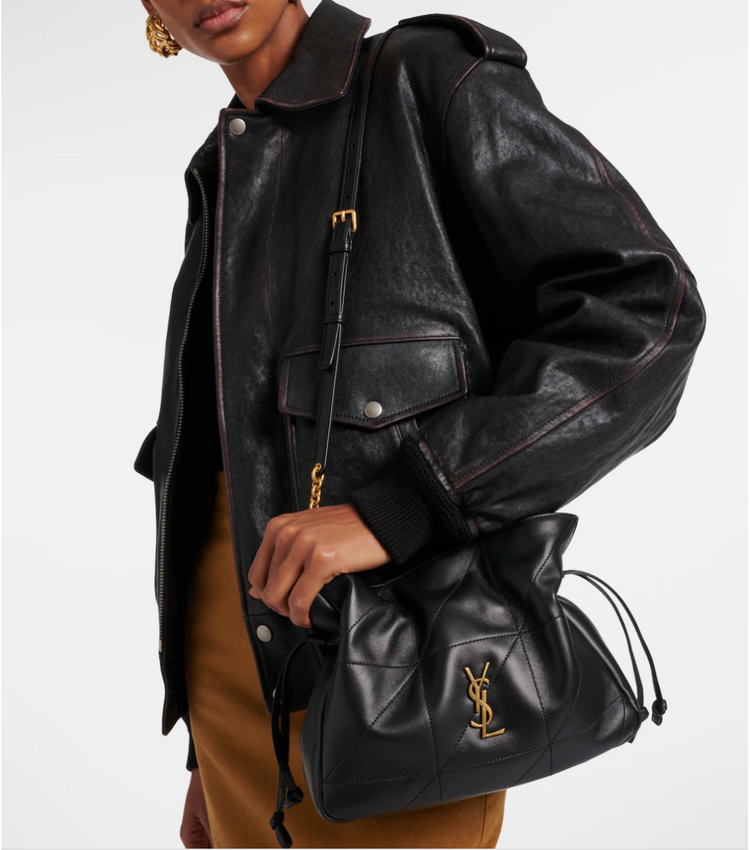 Person wearing a black leather jacket holding a black handbag with a YSL gold logo.