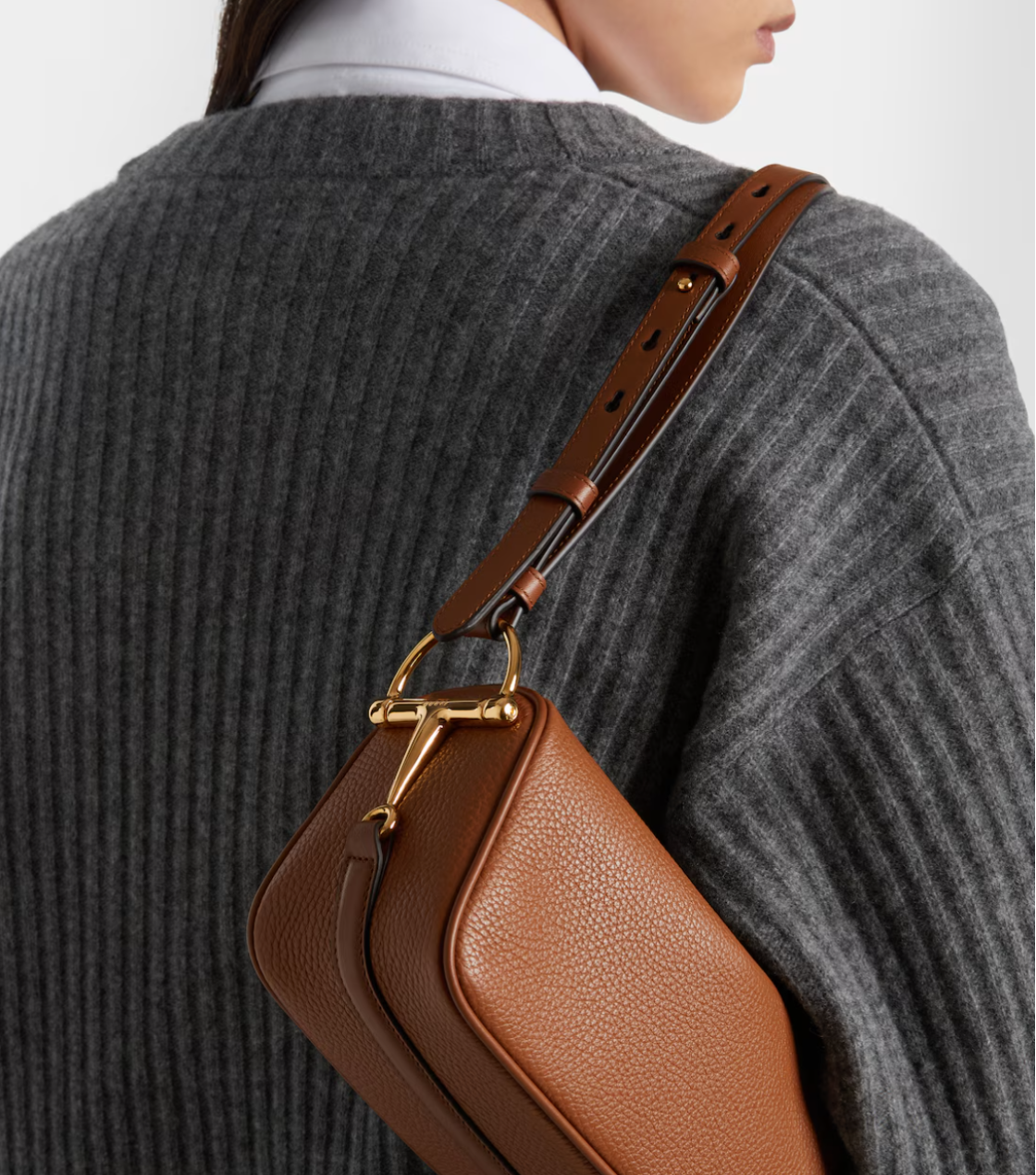 Brown leather handbag with gold hardware worn by a person in a gray sweater.