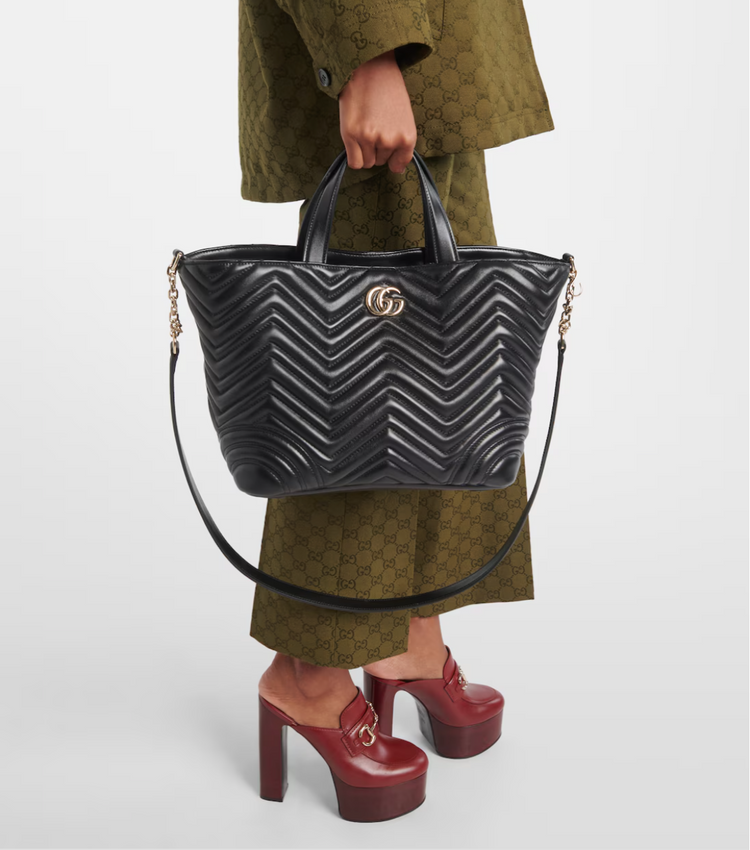 Person holding a black quilted handbag with a brand logo, wearing a green coat and red shoes.