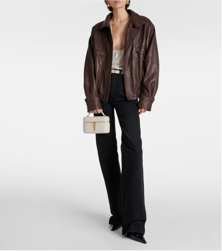 Person wearing a brown leather jacket and black pants holding a white handbag on a light gray background